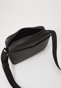 Black crossbody bag with a textured design, zip closure, and branded strap. Made from synthetic materials with a smooth finish.