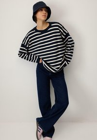 Pasiasty sweter w kolorze granatowym i kremowym, o oversize'owym kroju z długimi rękawami, zestawiony z ciemnoniebieskimi dżinsami w fasonie dzwonów oraz różowymi trampkami. Dodatkowo granatowy kapelusz typu bucket.