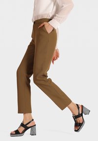 Pantaloni marroni su misura con una texture liscia e tasche laterali, abbinati a sandali neri con tacco a blocco decorato e cinturini alla caviglia.