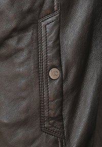 Bruine leren jas met een gladde textuur, gestikte details en een metalen knop met een logo. Close-up van het zakgebied.