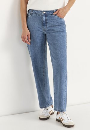 Persoon draagt lichtblauwe rechte jeans en witte sneakers met bruine strepen, staand tegen een witte achtergrond.