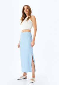 Lichtblauwe maxi-rok van textiel met zijsplitten, gecombineerd met een witte cropped tanktop en witte instappers met decoratieve banden.