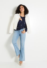 Chaqueta de denim blanca sobre un suéter azul marino con patrones de estrellas, combinada con jeans acampanados de un azul claro y tacones dorados. Atuendo simple y casual.