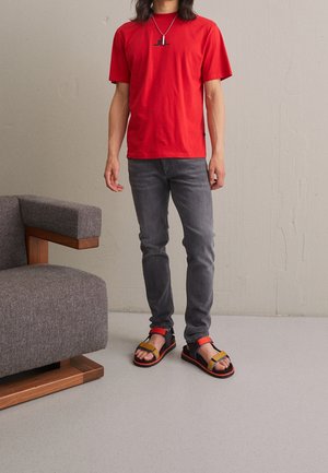 Eine Person steht neben einem grau gepolsterten Stuhl mit Holzakzenten und trägt ein rotes T-Shirt, graue Jeans, bunte Sandalen und eine Halskette.