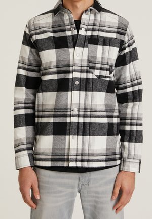 Homme portant une chemise à carreaux noire, blanche et grise avec poche poitrine et pantalon gris clair sur fond uni.