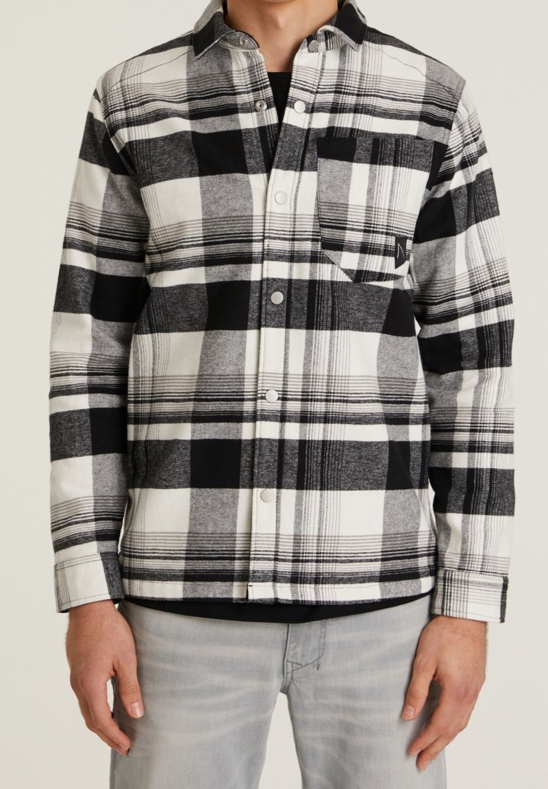 Homme portant une chemise à carreaux noire, blanche et grise avec poche poitrine et pantalon gris clair sur fond uni.