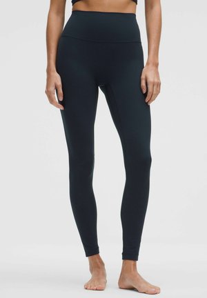 Kvinde iført sorte, højtaljede leggings i fuld længde, stående barfodet mod en ensfarvet grå baggrund.
