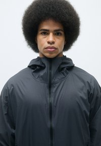 Veste zippée noire avec col montant et capuche, dotée d'une texture lisse et légère ainsi que de poches latérales. Le modèle a une coiffure afro.