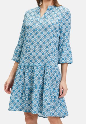 Femme portant une robe mi-longue bleue et blanche à motifs, avec des manches cloche trois-quarts et une jupe à volants.