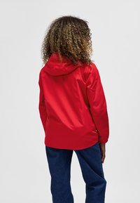 Red waterproof jacket with a hood, elastic cuffs, and a smooth texture, paired with denim pants. View shows the back of the jacket.