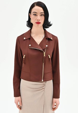 Donna con capelli neri corti indossa una giacca marrone con cerniera e una gonna beige chiaro arricciata, in piedi davanti a uno sfondo bianco semplice.