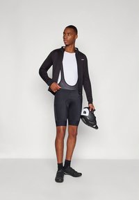 Svart sykkeljakke over en hvit sykkeltrøye, kombinert med svarte polstrede sykkelshorts. Svarte sykkelsko og en svart hjelm holdes i én hånd.