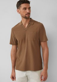 Brązowa koszulka polo z krótkim rękawem, z dekoltem w kształcie litery V i detalem kieszeniowym. Teksturowana tkanina i luźny krój. Stylizowana z jasnymi spodniami.