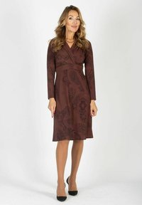 Brązowa sukienka z długimi rękawami, zapinana w talii, w wzór paisley, dopasowana w talii, sięgająca do kolan. Modelka nosi cieliste rajstopy i czarne obcasy.