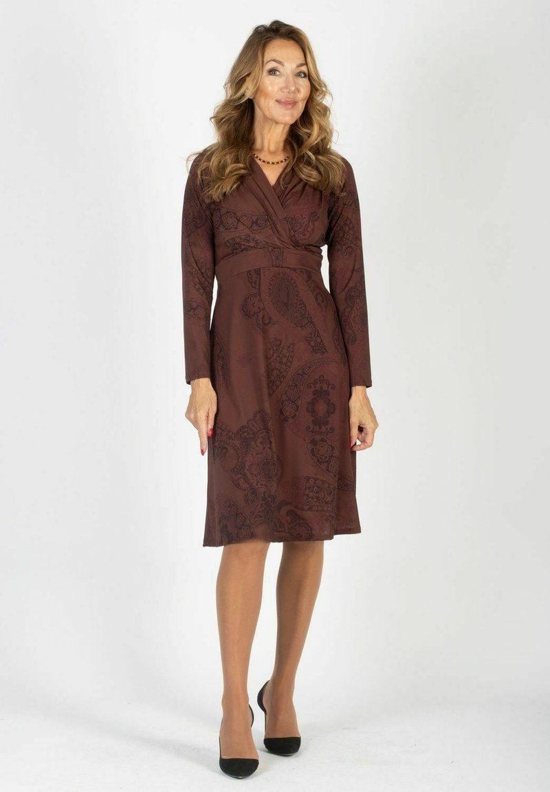 Brązowa sukienka z długimi rękawami, zapinana w talii, w wzór paisley, dopasowana w talii, sięgająca do kolan. Modelka nosi cieliste rajstopy i czarne obcasy.