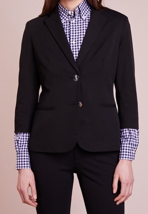 Vrouw draagt een aansluitende zwarte blazer over een blauw-wit geruit overhemd met knopen, met bijpassende geruite manchetten die onder de blazermouwen zichtbaar zijn.