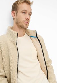 Beige Fleecejacke mit hohem Kragen, schwarzem Reißverschluss und blauen Akzenten. Trägt darunter ein helles Creme-T-Shirt mit Brusttasche.