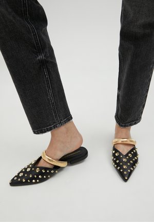 Černé mule s špičatou špičkou a zlatými kovovými doplňky, zdobené kovovými nýty podél horní části a robustním popruhem na patě.