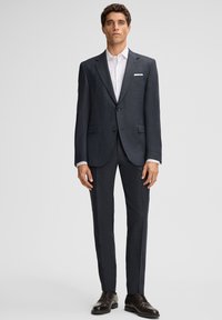 Ein Mann, der in einem dunkelgrauen Anzug mit einem weißen Hemd, schwarzen Leder-Schuhen und einem weißen Taschentuch vor einem schlichten Hintergrund steht.