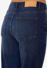 Tamsiai mėlyni džinsai iš džinsinio audinio, prigludusio dizaino, matomomis siūlėmis ir dviem užpakalinėmis kišenėmis. Juosmens juostoje siuvinėta ruda odinė etiketė.