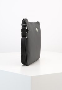 Borsa sintetica nera con finitura testurizzata, chiusura a zip e tracolla regolabile. Presenta un piccolo logo verde sul lato anteriore.