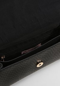 Černá texturovaná kabelka s vnitřní kapsou na zip a zlatým zacvakávacím uzávěrem, s viditelnou značkovou etiketou. Podšita černou látkou.