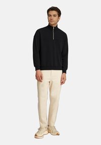 Felpa nera con mezza cerniera e collo strutturato, indossata con pantaloni color crema e sneakers chiare, caratterizzata da dettagli testurizzati.