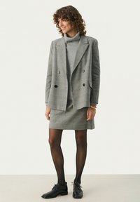 Gråternet blazer med dobbeltradede knapper, parret med en grå turtleneck og matchende nederdel. Sorte strømpebukser og sko fuldender looket.