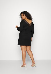 Vestido preto com mangas longas, detalhes em renda na gola, bainha em forma de ondas e um ajuste solto. Modelo a usar sandálias de salto bege.