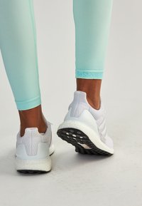 Białe buty sportowe z teksturowaną siateczką, amortyzowaną podeszwą i czarnym bieżnikiem. Jasnoniebieskie legginsy z brandingiem "BeShaped" na końcu.