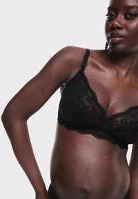 Sort blonde-bh med skarpe kanter, blød tekstur og justerbare stropper, båret af en person, der viser en rund mave.