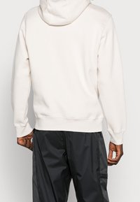 Ljusbeige hoodie med avslappnad passform, med dragsnodd i huvan, ribbade mudd och nederkant, som bärs tillsammans med mörka, lätta cargobyxor.