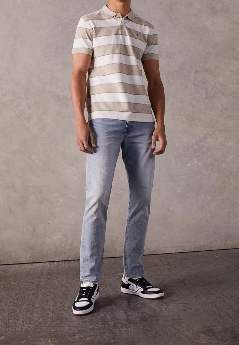 Pruhované krátké polo tričko v krémové a béžové barvě, v kombinaci se světle modrými džíny a černo-bílými teniskami, stojící na texturované podlaze.