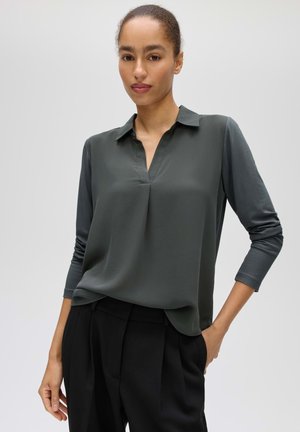 Donna con capelli tirati indietro che indossa una camicetta grigio scuro a maniche lunghe e pantaloni neri, in piedi con una mano in tasca.