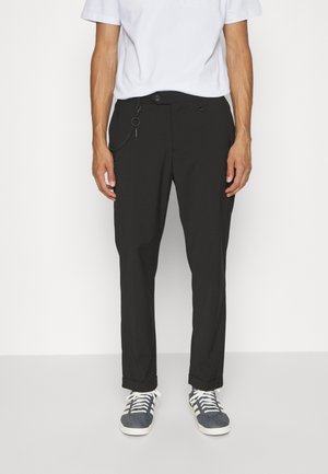 Homme portant un pantalon noir avec une chaîne latérale, un t-shirt blanc et des baskets grises, debout devant un fond blanc uni.
