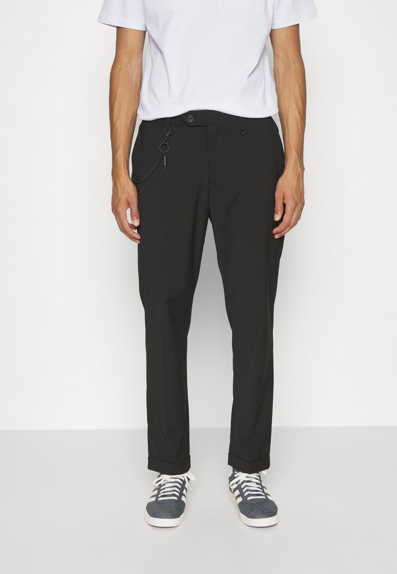 Homme portant un pantalon noir avec une chaîne latérale, un t-shirt blanc et des baskets grises, debout devant un fond blanc uni.