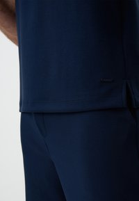 Granatowa bawełniana koszulka z luźnym krojem, z bocznymi rozcięciami na dole i małą metką z logo przy szwie. Gładka tekstura i prosty design.