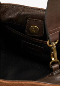 Interior de un bolso de cuero marrón, con un forro de tela oscura, cierre de imán y un pequeño bolsillo con una etiqueta de logo. Herrajes en tono dorado.