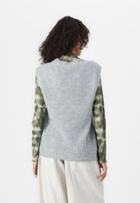 Grijze gebreide mouwloze trui met ribbelstructuur, gedragen over een groen tie-dye langemouwen top. Lichtgekleurde broek maakt de outfit af.