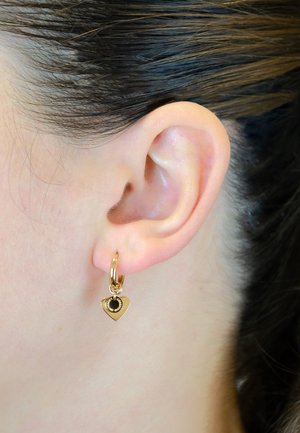 Des boucles d'oreilles en or en forme de boucle présentent un charme en forme de cœur avec un détail central noir. Le design comprend des bords arrondis et une finition lisse.
