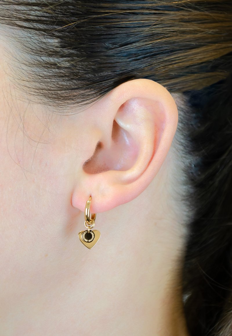 Des boucles d'oreilles en or en forme de boucle présentent un charme en forme de cœur avec un détail central noir. Le design comprend des bords arrondis et une finition lisse.