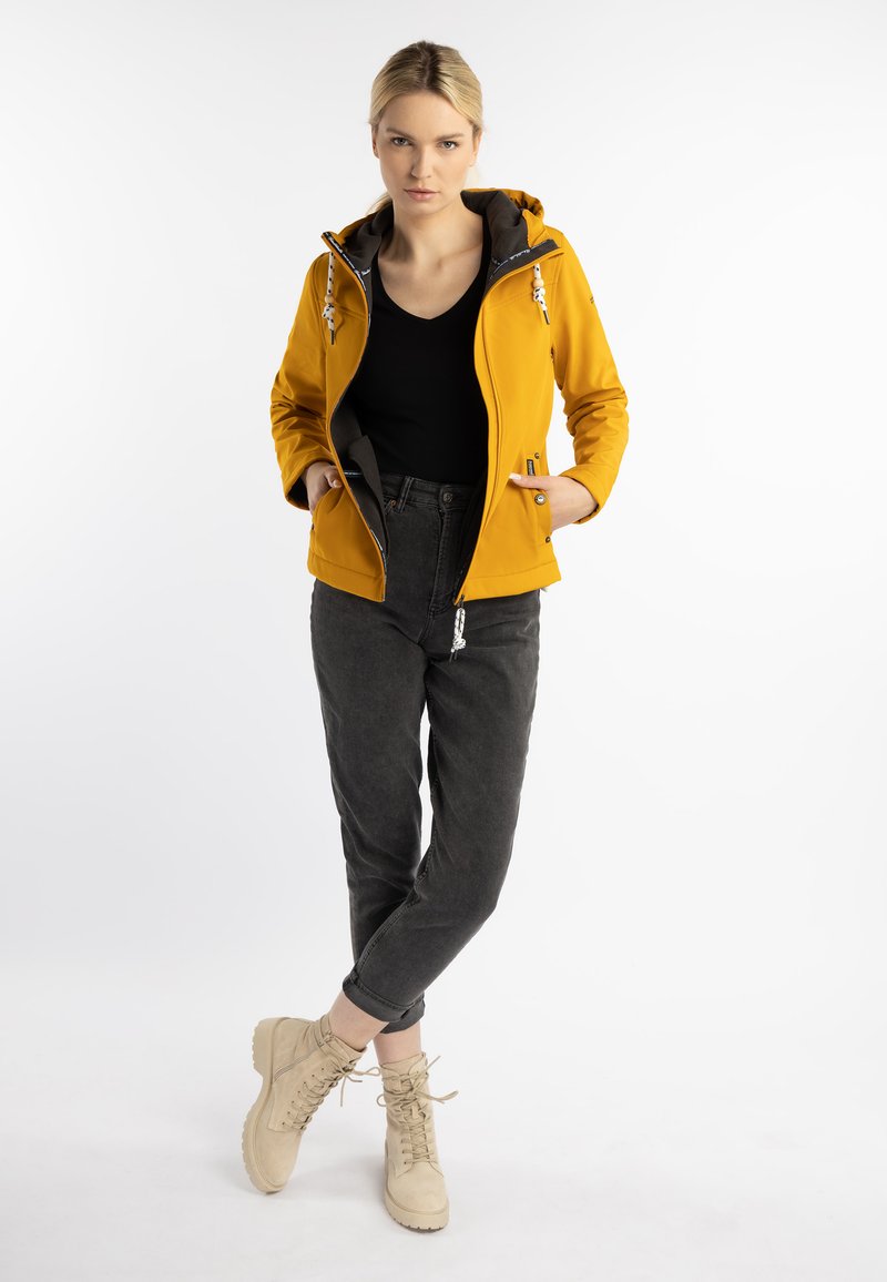 Chaqueta softshell amarilla con capucha gris, bolsillos con cremallera y detalles de ajuste, combinada con pantalones gris oscuro y botas beige.