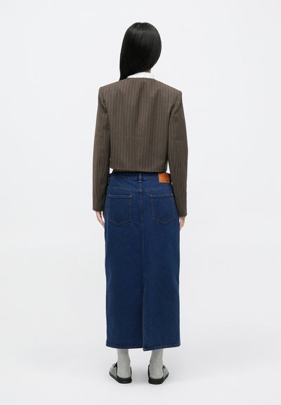 Barna csíkos rövid blézer, kék farmer hosszú szoknya hasítékkal, világosbarna folt a hát alsó részén, szürke zokni és fekete slip-on cipő.