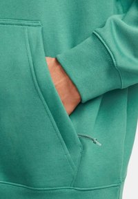 Grön hoodie med känguruficka, ribbade ärmslut och en slät yta. Inkluderar en dragsko närmast fickan.