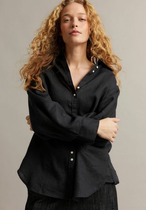 Frau mit lockigen blonden Haaren, die ein locker sitzendes schwarzes Hemd mit Knöpfen trägt und mit verschränkten Armen vor einem schlichten hellen Hintergrund steht.