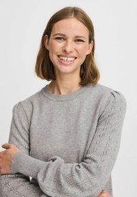 Harmaa neulepaita, jossa on pitkät hihat; siinä on pyöreä kaula-aukko ja koristeelliset reiät hihoissa; pehmeä rakenne ja hieman puffatut olkapäät.