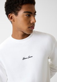 Biała koszula z długim rękawem, z okrągłym dekoltem i prążkowaną fakturą, z czarnym haftem "Guess Jeans" na piersi.