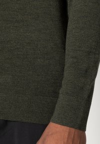 Primo piano del braccio di una persona che indossa un maglione lavorato a maglia verde scuro con polsini e orlo a coste, abbinato a pantaloni neri.