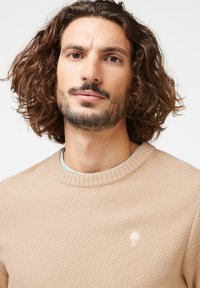 Beige strukturierter Pullover mit rundem Halsausschnitt und gerippten Abschlüssen. Verfügt über ein dezentes Logo auf der Brust, das über einem hellblauen Hemd getragen wird.