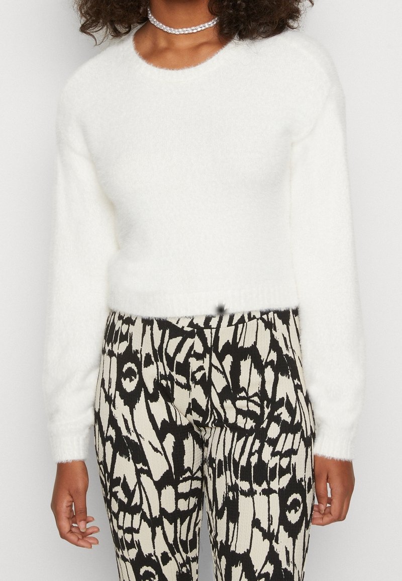 Femme portant un pull à manches longues blanc duveteux, un pantalon noir et blanc à motif, et un collier transparent à perles, debout devant un fond uni.